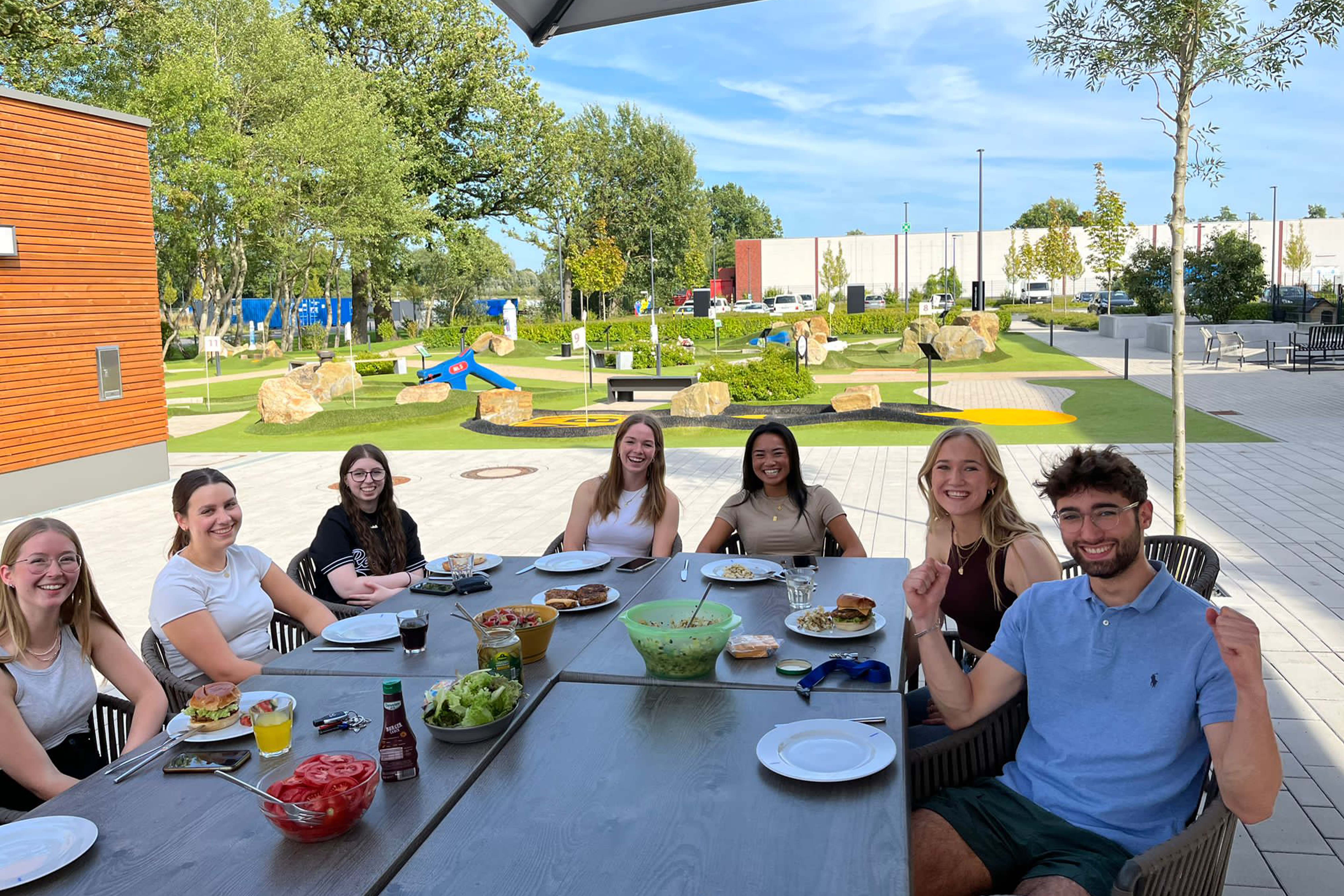 Un grupo de jóvenes se sienta alrededor de una mesa puesta, riendo, con un campo de minigolf al fondo.