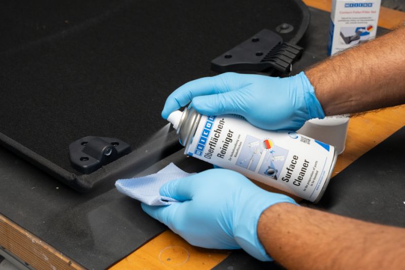 Manos con guantes azules pulverizan Surface Cleaner de un aerosol sobre el borde de una pieza plástica negra mientras limpian con un paño
